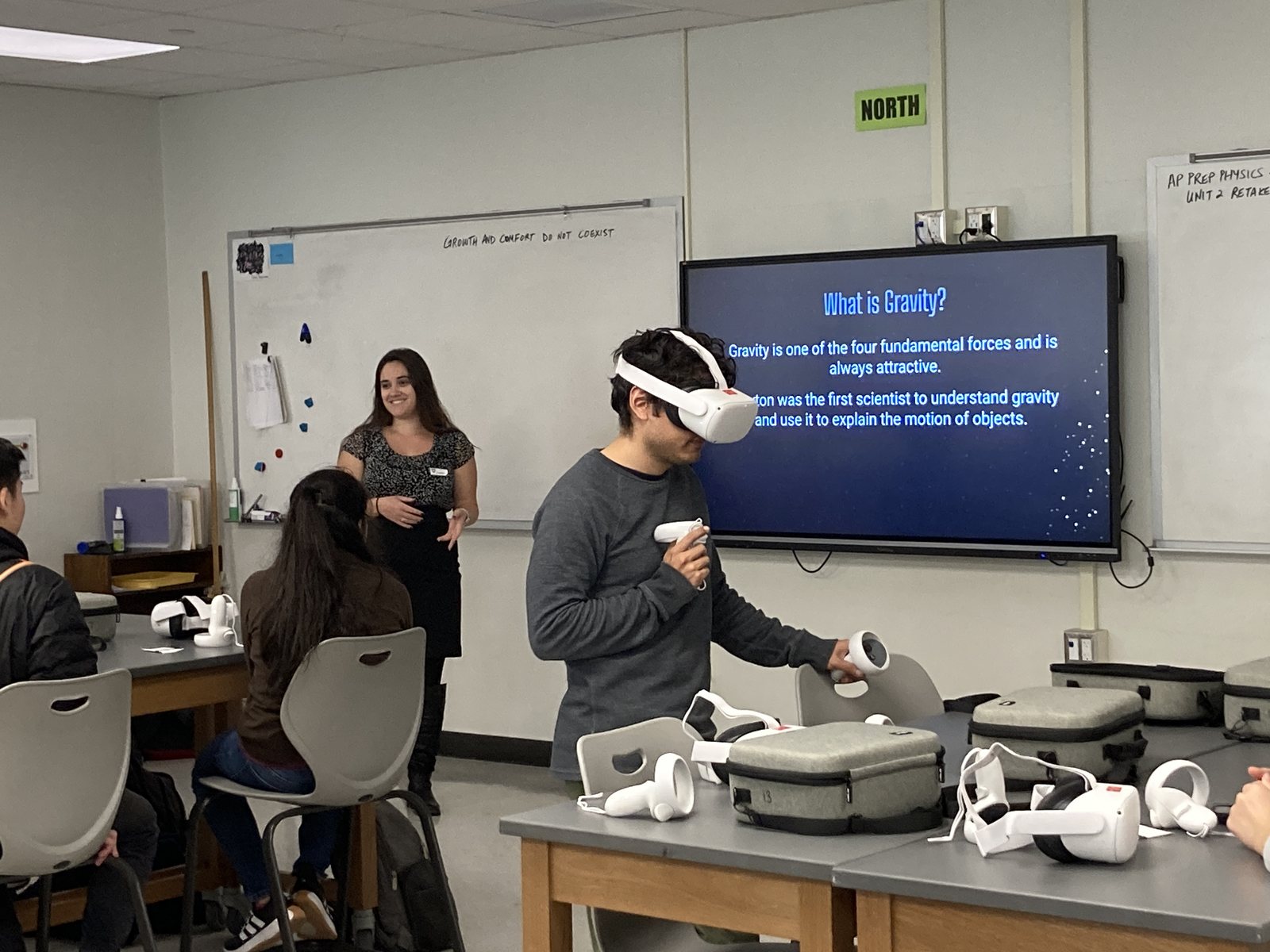 A high-school classroom: a student wearing a virtual-reality headset holds the controllers while Kristen speaks to the class, with a slide reading What is Gravity? on the screen behind and more headsets on the desks